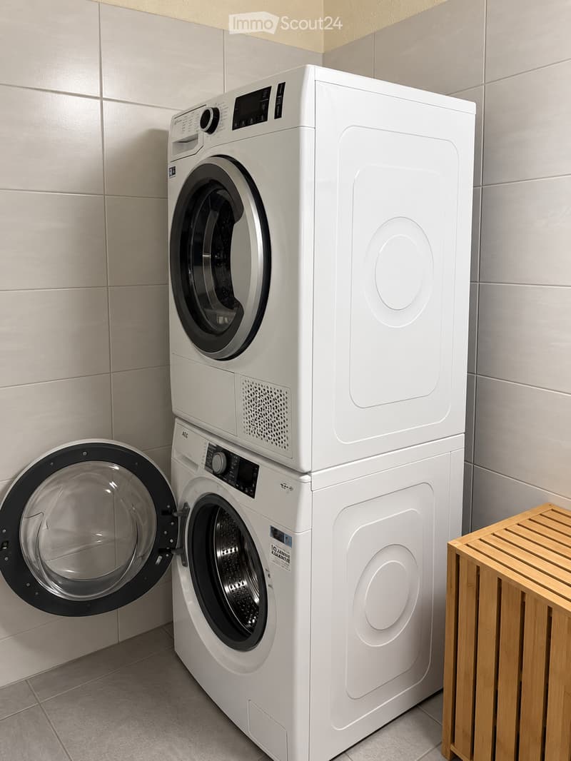 A modern bathroom with a white washing machine and dryer stacked vertically, featuring a wooden shelf to the right.