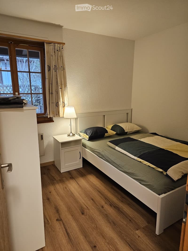Cozy bedroom with a single bed, nightstand, lamp, and window with curtains. Wooden flooring and light-colored walls.