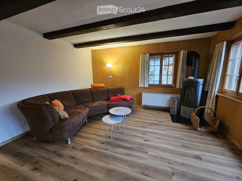 Cozy living room with wooden flooring, exposed wooden beams, a brown sectional sofa, a small white coffee table, a white stool, a fireplace, and a floor lamp. There are also two wicker baskets near the fireplace.