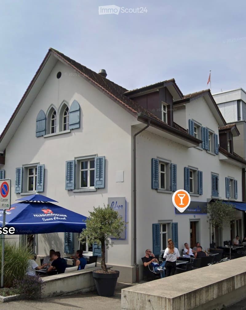 Outdoor seating area of a restaurant with tables, chairs, and umbrellas, featuring a potted tree and a sign for "Blue" on the building.