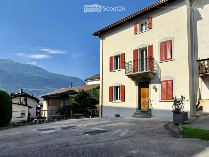 Two-story house with light yellow walls, red shutters, and a balcony overlooking a driveway. The house features a wooden front door and is situated in a scenic area with mountains in the background.