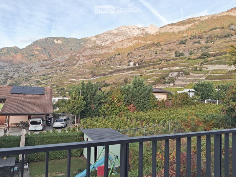 Balcony overlooking a scenic valley with terraced vineyards and mountains, featuring a small patio area with a table, chairs, and an umbrella, as well as a garage with parked vehicles and bicycles.