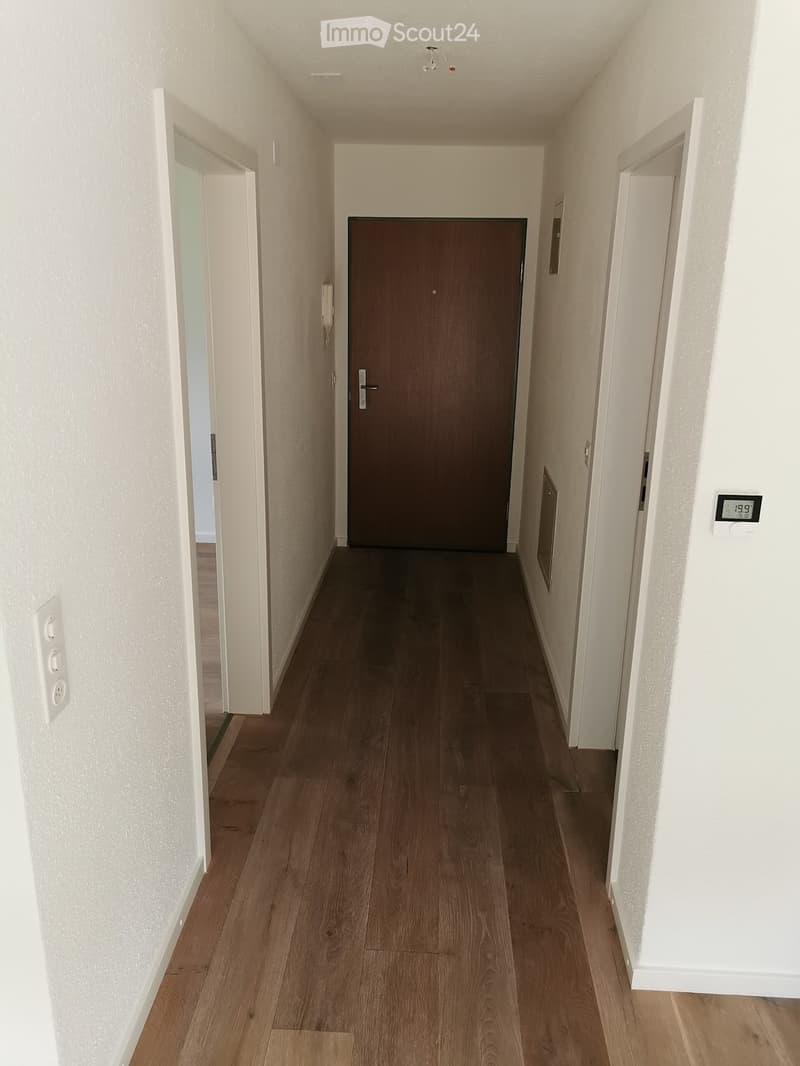 A clean, well-lit hallway with wooden flooring, white walls, and a central closed door. The hallway has a thermostat on the right wall and a light switch on the left wall.