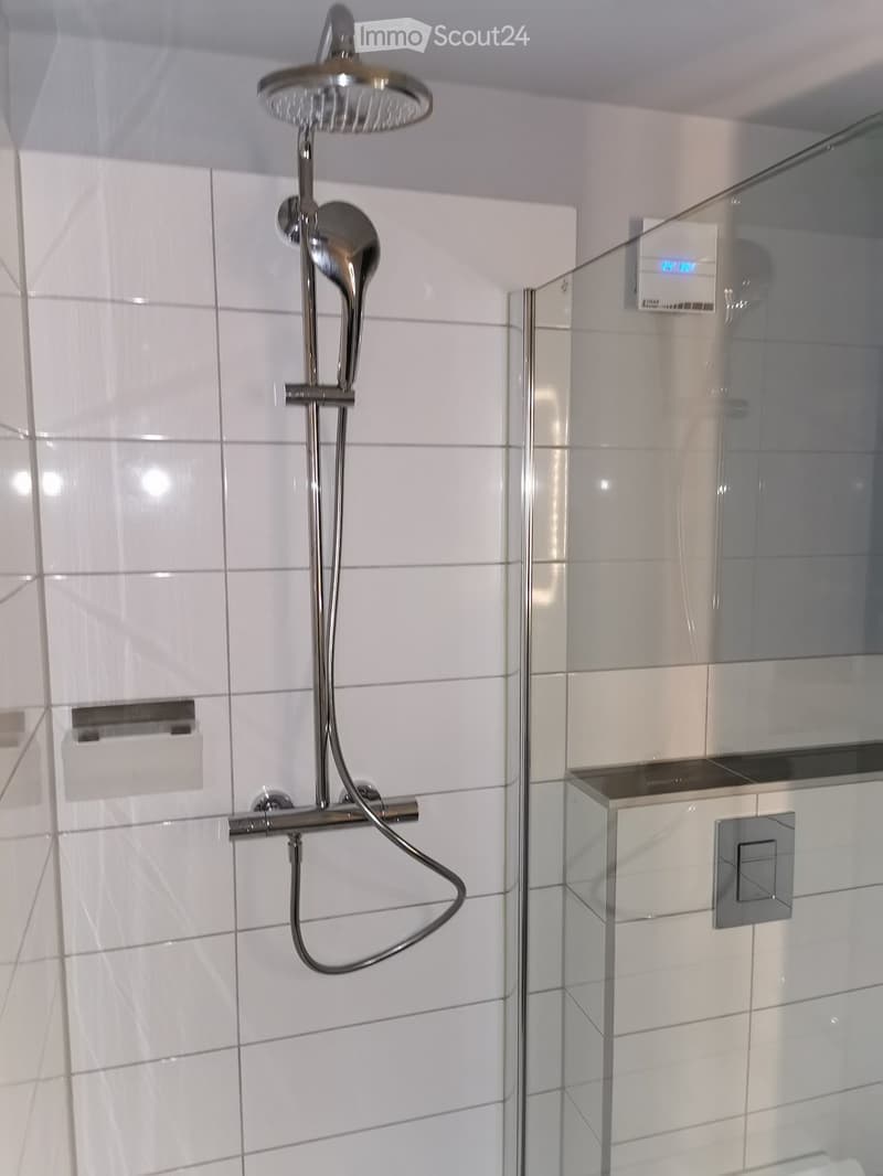 Modern bathroom shower area with a chrome showerhead, hose, and glass enclosure. White tiled walls and a control panel are visible.
