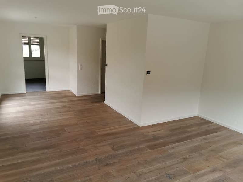 Empty room with light wooden flooring and white walls, featuring a doorway leading to another room with dark flooring.