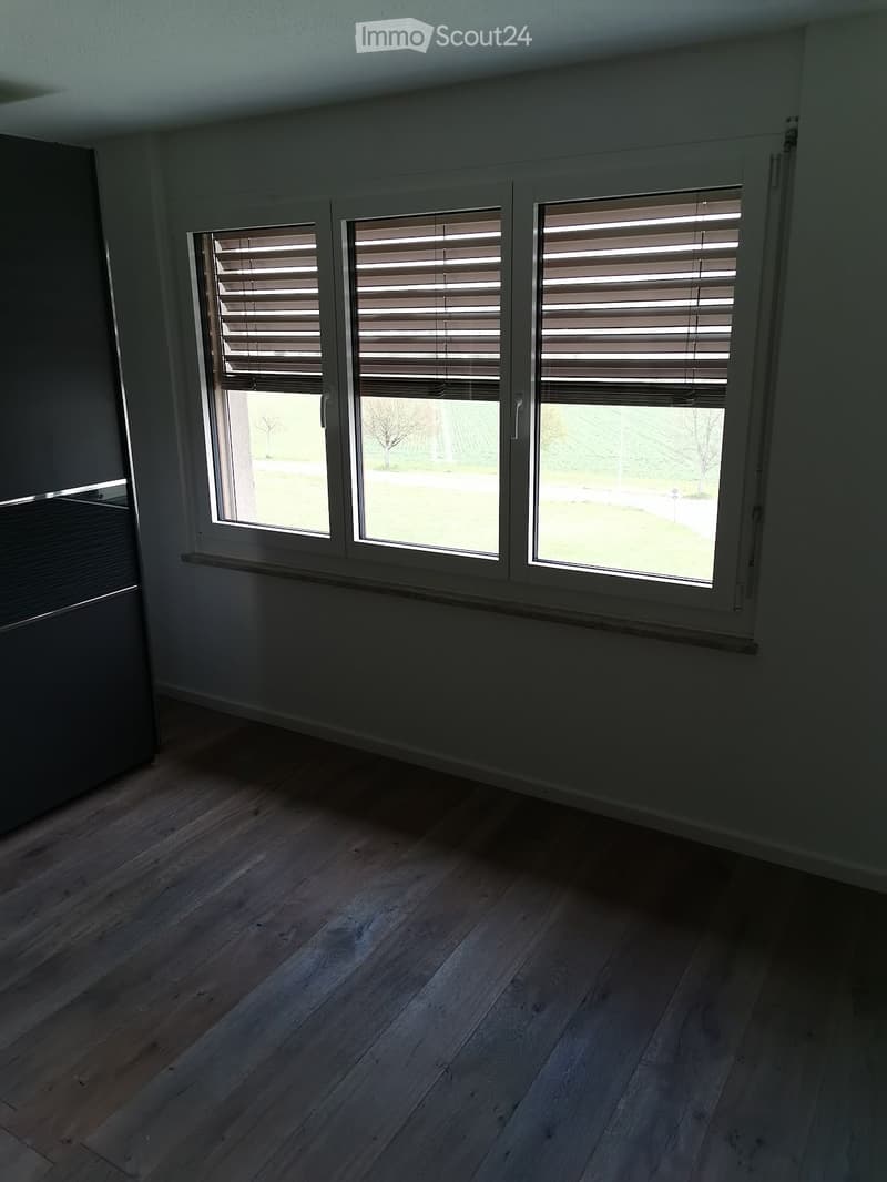 Empty room with wooden flooring, large windows with blinds, and a radiator below the windows.