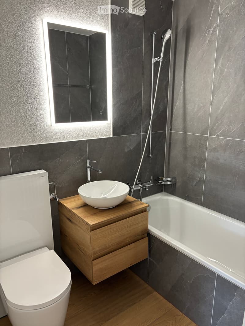 Bathroom with grey tiles, white sink, white bathtub, white toilet, wooden countertop, silver faucet, mirror with lighting