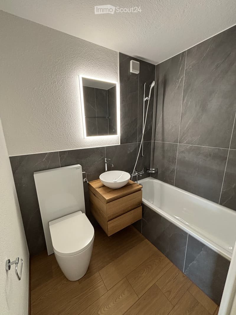 modern bathroom with bathtub, sink, toilet, illuminated mirror, white walls, wooden floor