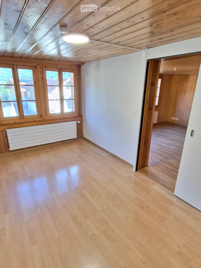 Empty room with light wooden flooring, white walls, large windows, and a visible radiator. The room has a wooden ceiling and a doorway leading to another room.