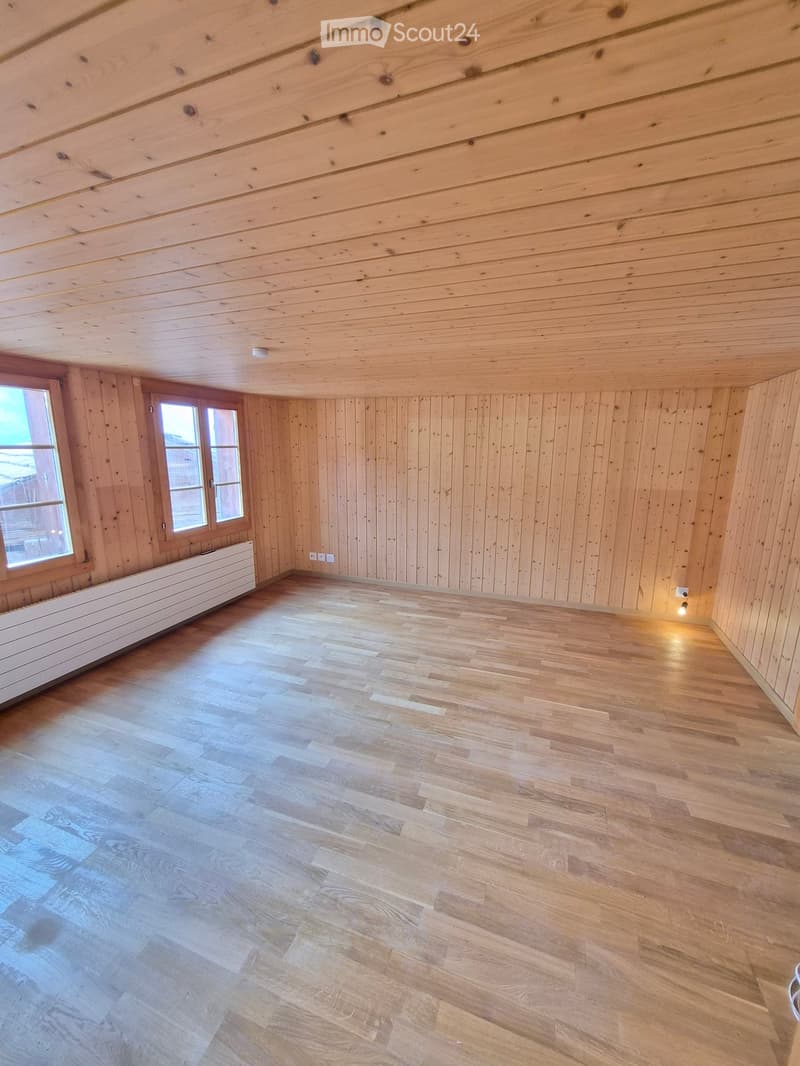 Empty room with wooden walls, ceiling, and floor, featuring two windows and a radiator on one wall.