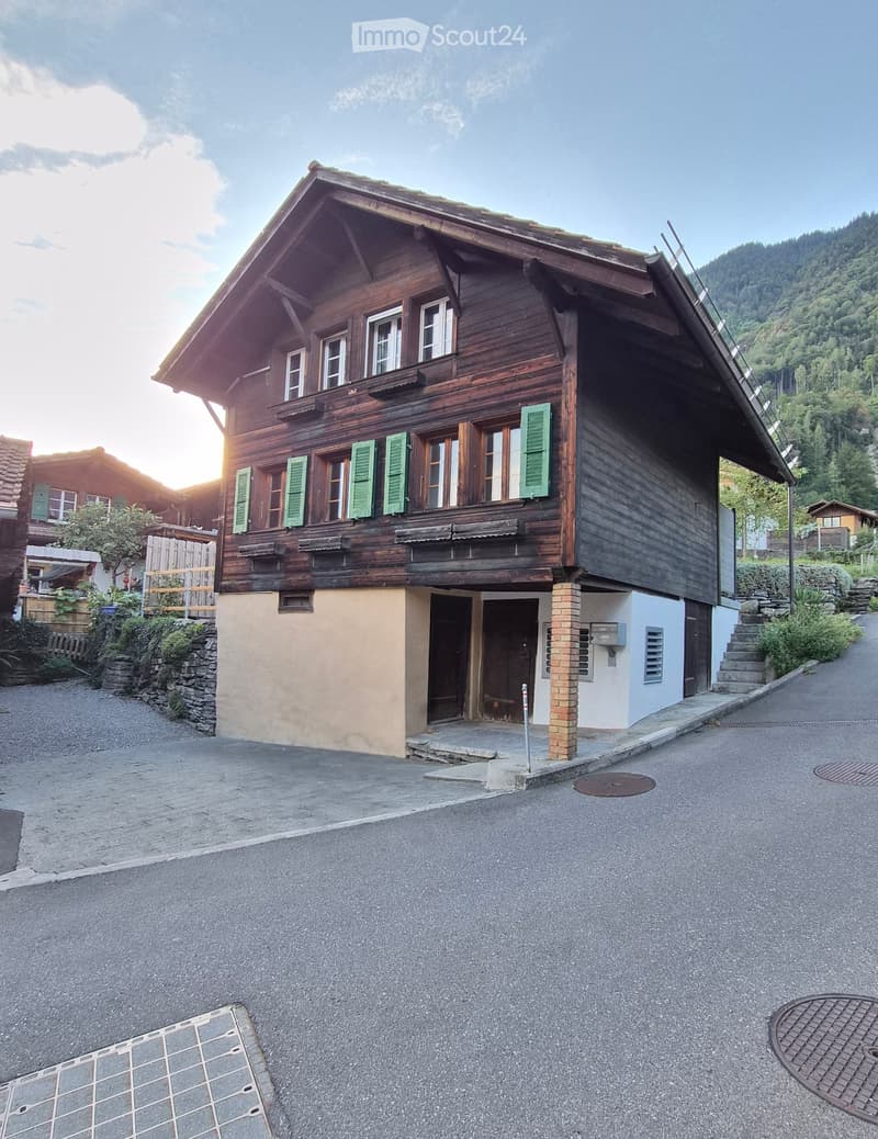 Traditional wooden house with green shutters, stone base, and a sloped roof, situated in a rural area with a gravel driveway and surrounding greenery.