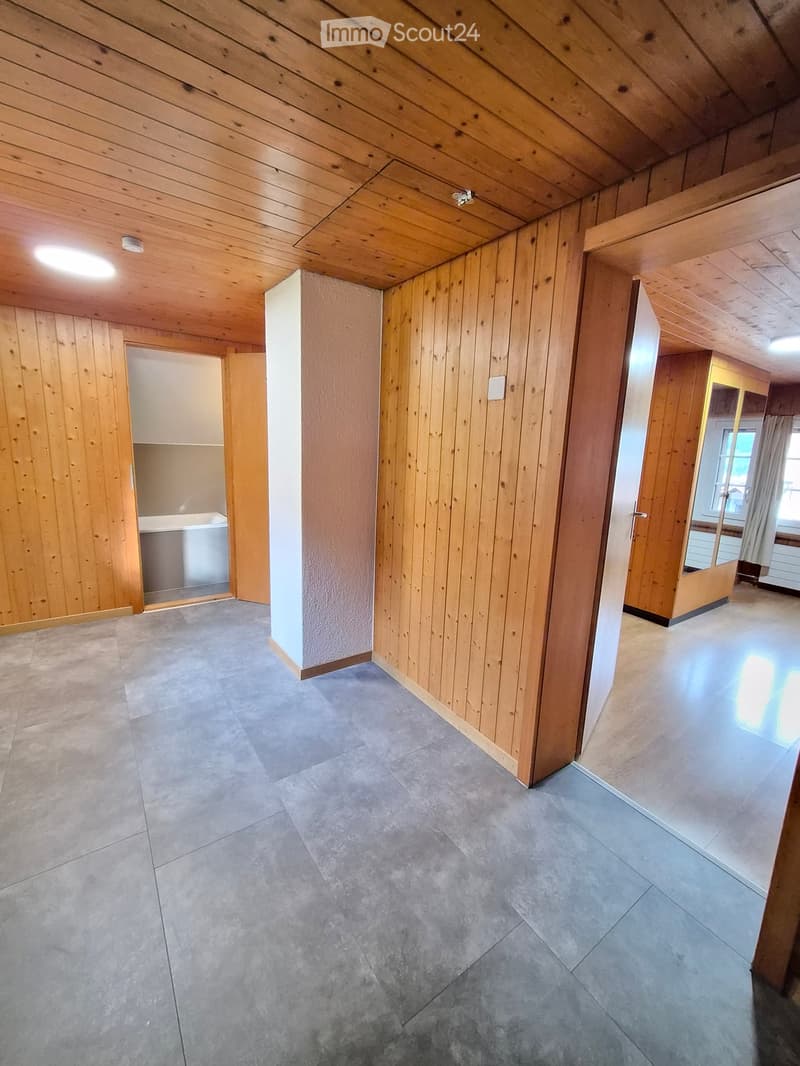 Empty room with wooden walls, exposed wooden ceiling beams, and grey tiled floor. No furniture or equipment visible.