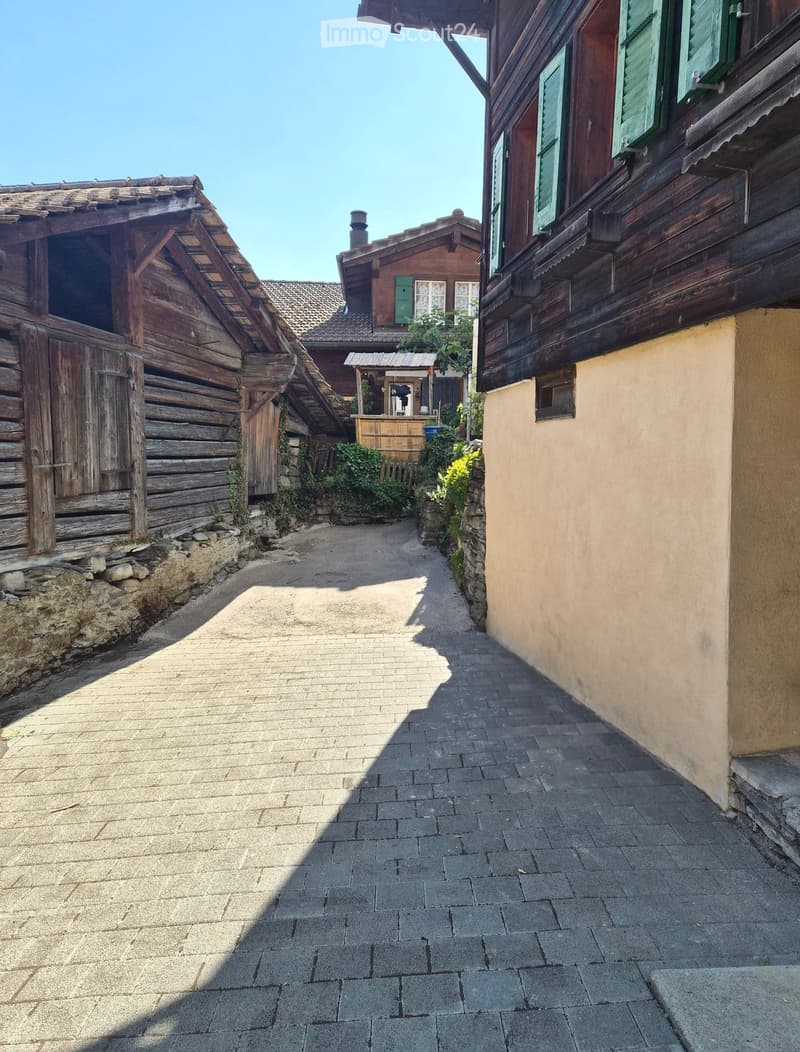 A narrow, cobblestone pathway flanked by traditional wooden houses with green shutters and stone walls, leading to a small wooden structure in the distance.