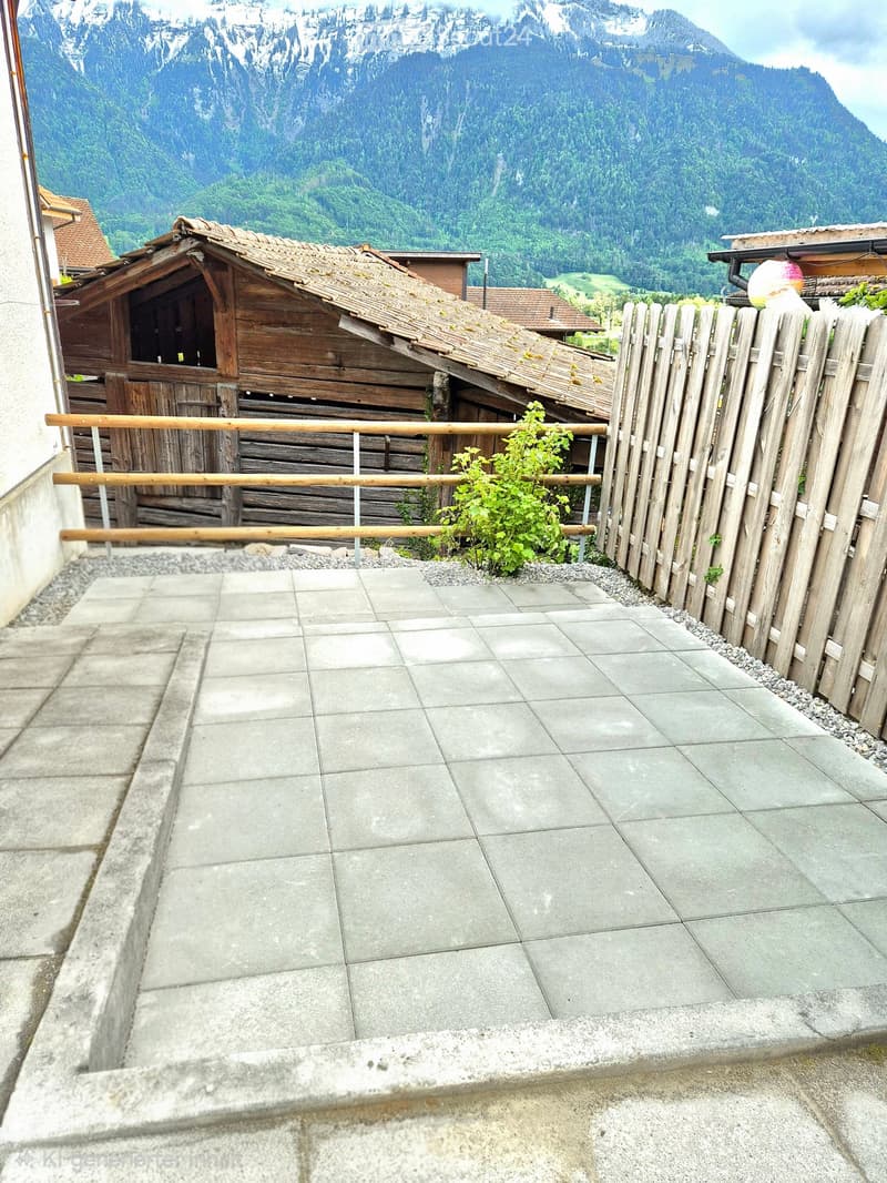 Balcony with tiled concrete floor, wooden railing, small green plant, and wooden fence, overlooking a scenic mountain landscape.