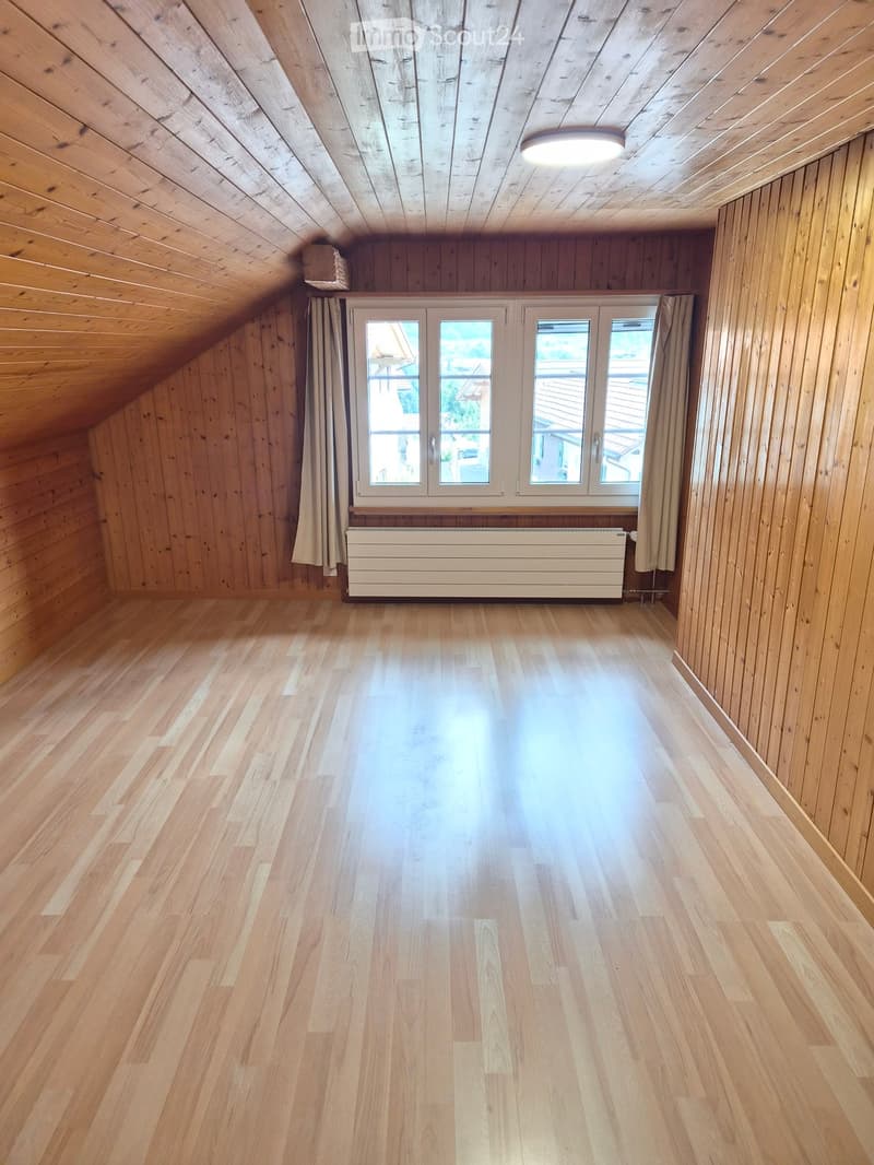 Empty attic room with wooden walls, wooden floor, large windows, and a radiator. No furniture or equipment present.