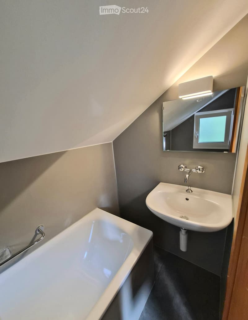 Modern bathroom with a bathtub, sink, and mirror. The room features a sloped ceiling and built-in lighting above the mirror.