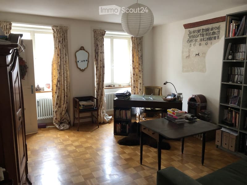 A well-lit room with wooden flooring, large windows with curtains, a sofa, a table, a bookshelf, a mirror, a lamp, and a radiator.