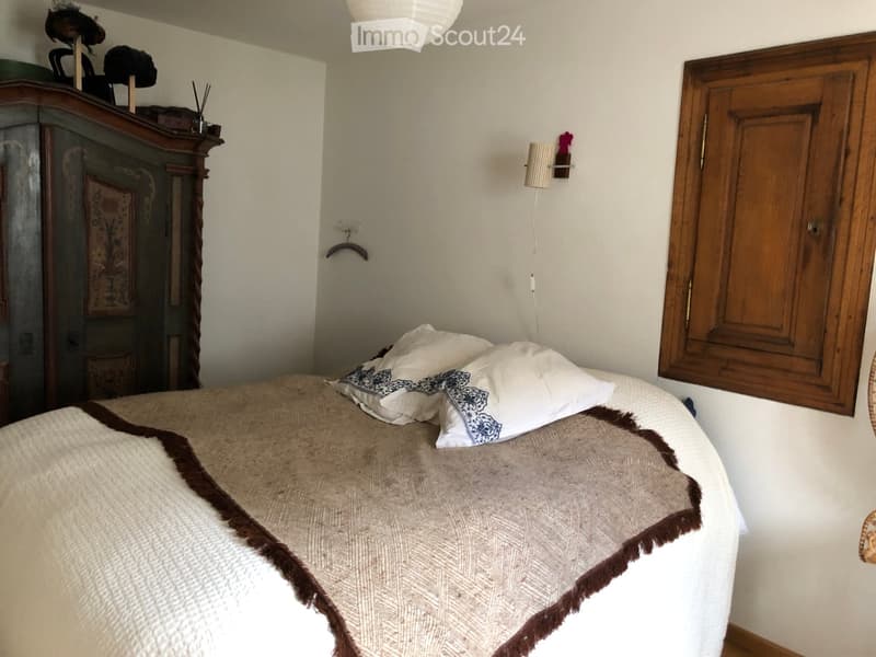 Cozy bedroom with a large bed, patterned pillows, a textured blanket, an antique wardrobe, and a wall-mounted lamp.