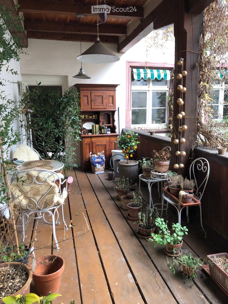 Wooden balcony with a hanging light fixture, a small table and chair, a wooden cabinet, and numerous potted plants.