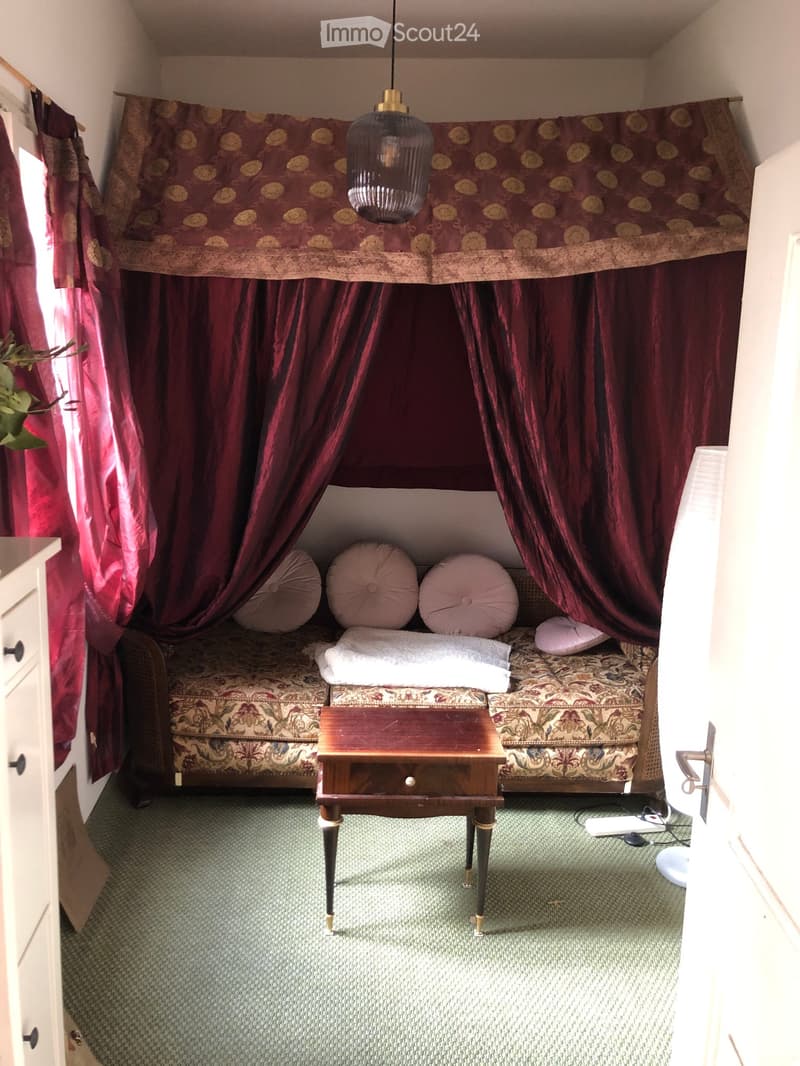 A cozy bedroom with a patterned sofa, decorative pillows, a small wooden table, and rich red curtains.