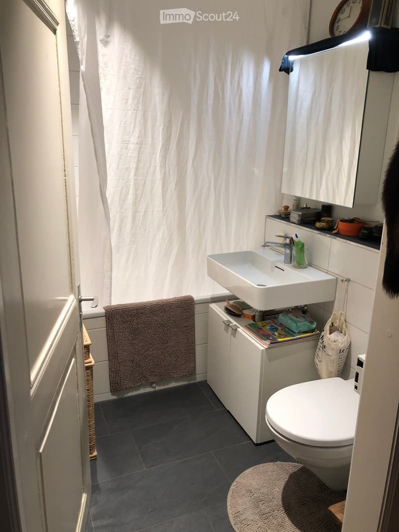 Modern bathroom with dark tiled floor, white sink, toilet, mirror, and a towel hanging on the wall. A shower curtain is visible, and a small basket and stool are placed near the door.