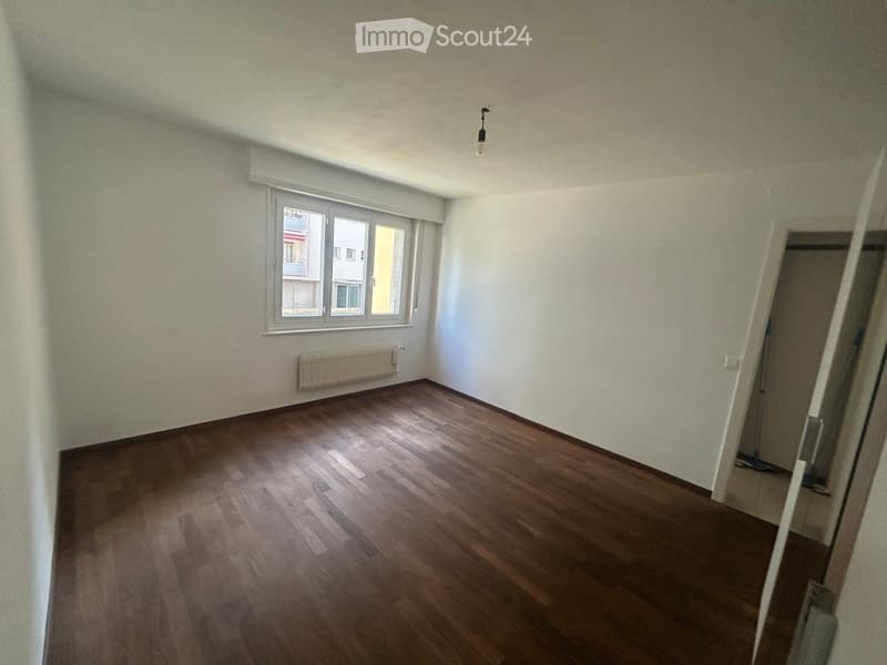 Empty room with wooden flooring, white walls, a window with a view, and a radiator below the window.