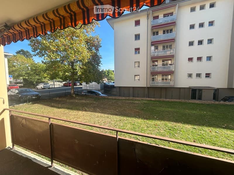 Balcony overlooking a grassy area with a multi-story building in the background, featuring balconies and windows, and a parking area with several cars.