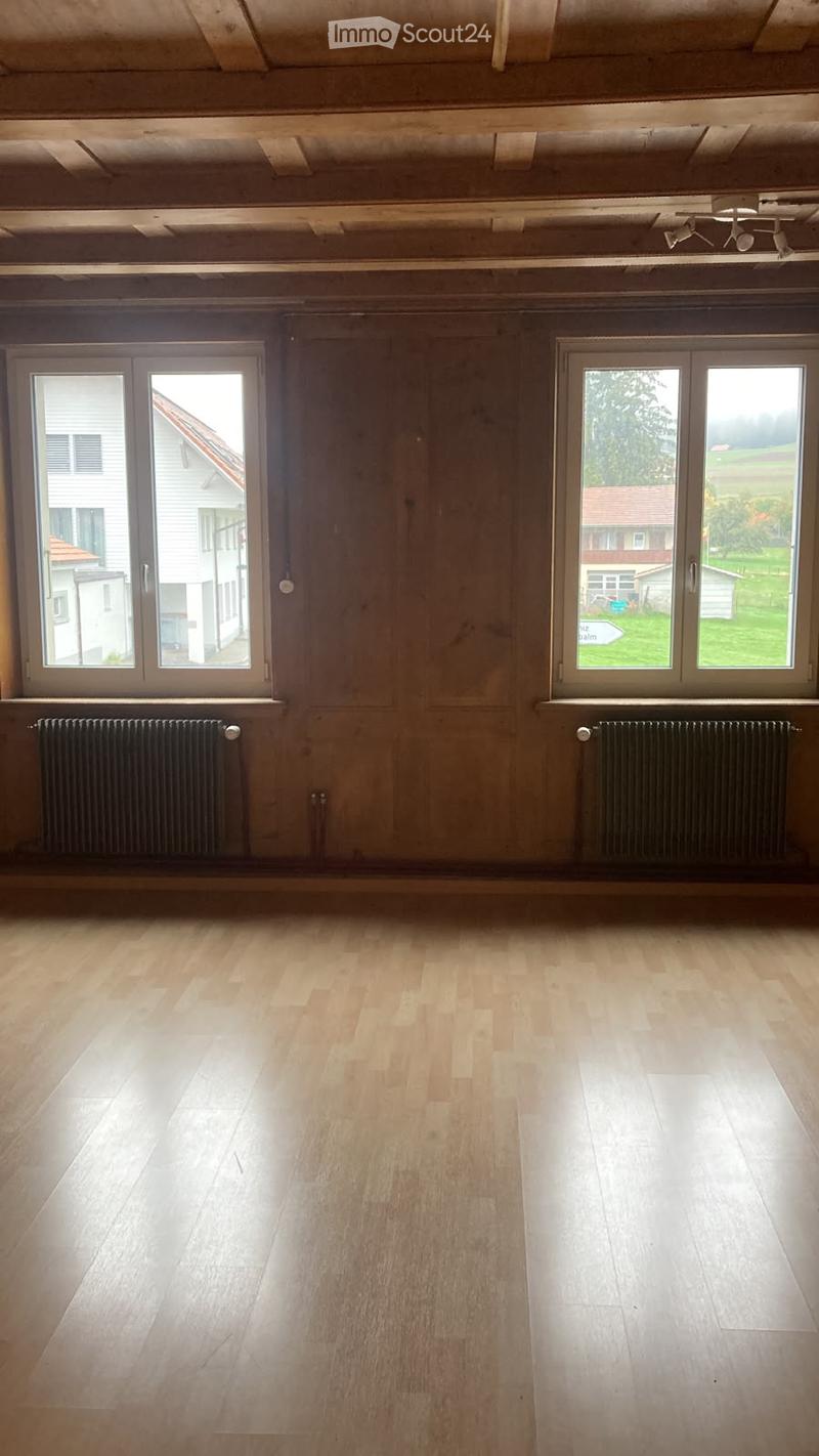 Empty room with wooden flooring, exposed wooden beams on the ceiling, large windows, and radiators along the walls.