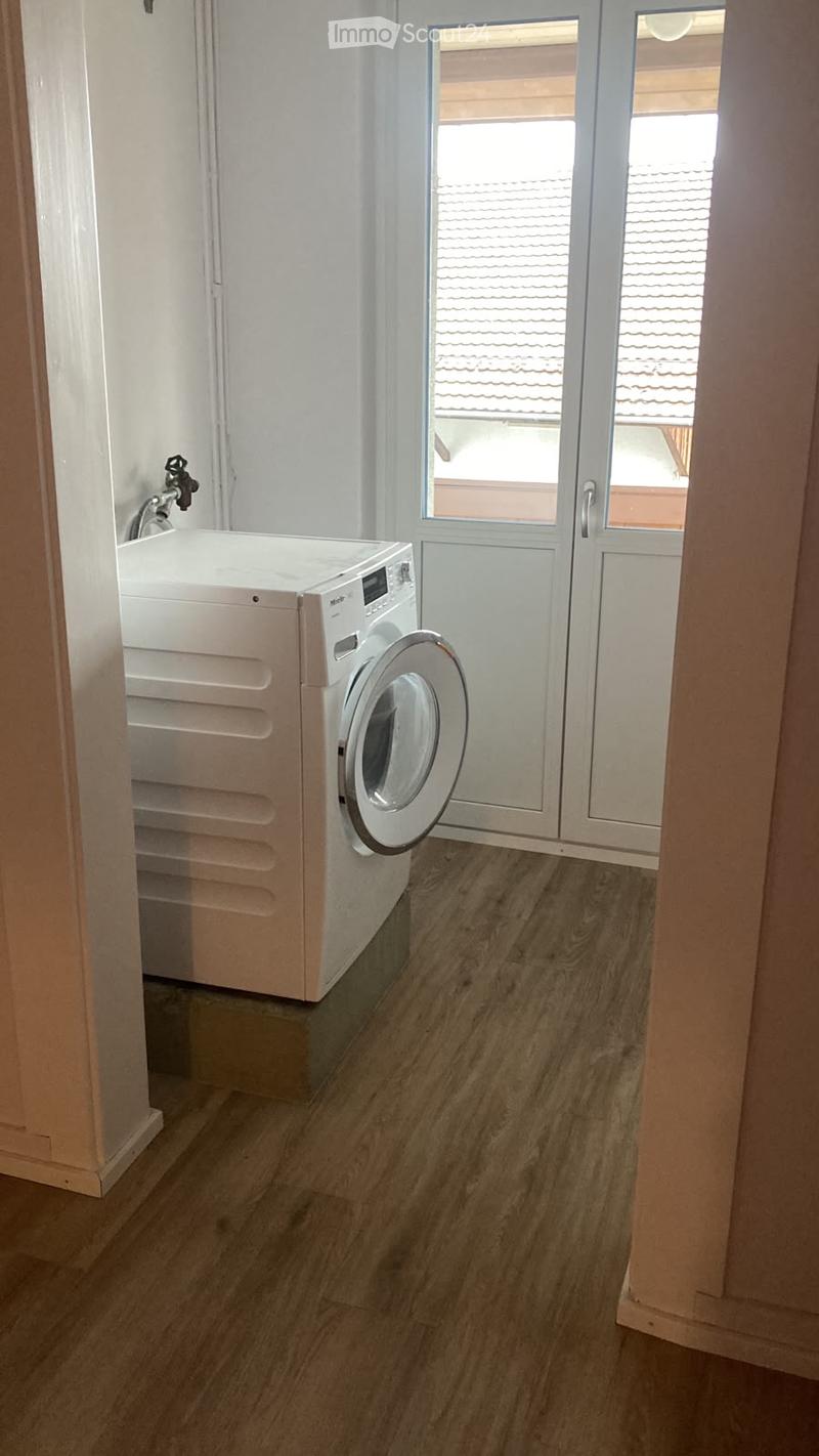 A small, well-lit room with wooden flooring and a white washing machine positioned against the wall. The room has a door leading to an outdoor area with a tiled roof visible through the glass.