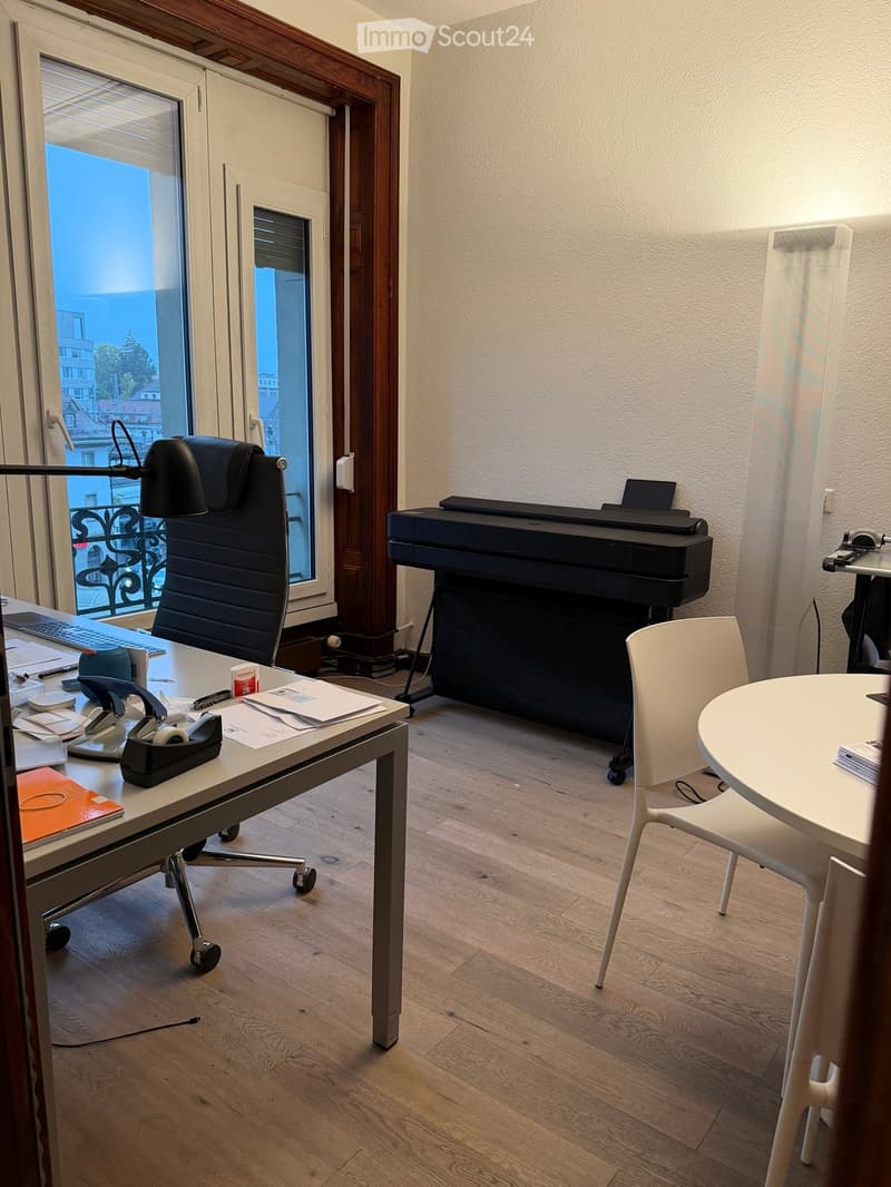 Office space with a desk, office chair, printer, round table, and white chair. The room has wooden flooring and large windows with a view of the city.