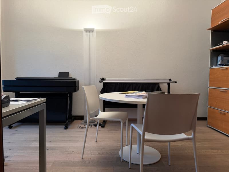 A modern, furnished room with a round white table, two white chairs, a printer, and a wooden cabinet with drawers.