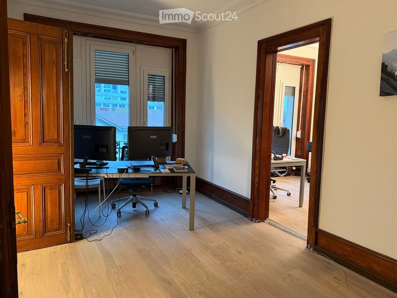 Office space with wooden flooring, two desks, chairs, and computer screens.