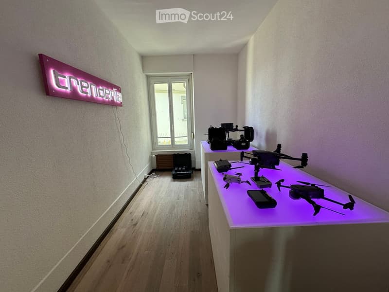 A small, well-lit room with wooden flooring and white walls, featuring a display table with drones and a cellphone on a purple-lit surface, along with a sign that reads "trendsia" on the wall.