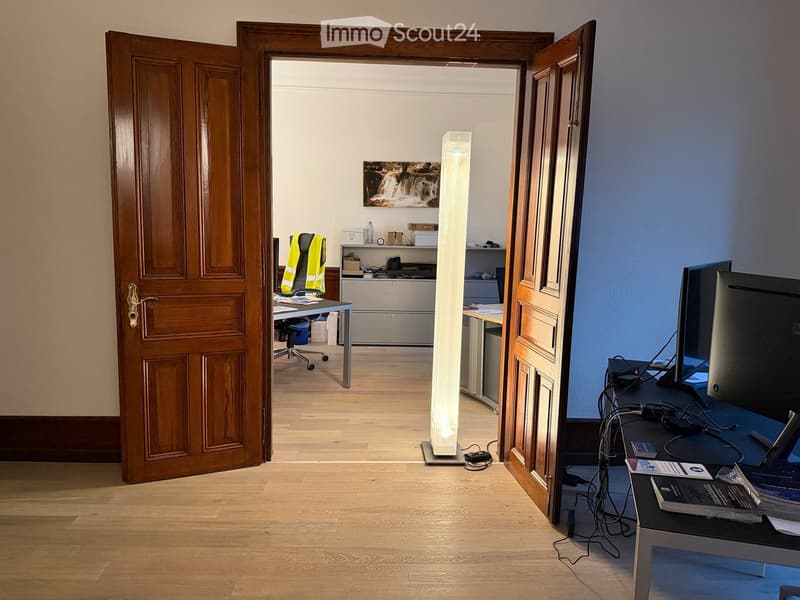 Office space with wooden flooring, open doorway leading to another room, a tall lamp, a chair, a table, a screen, and a cabinet.