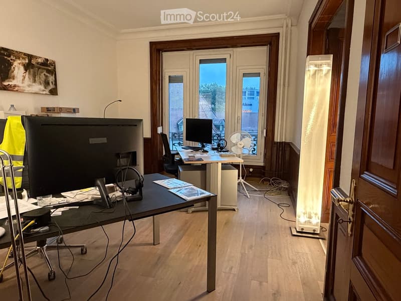 Office space with wooden flooring, large window, desk with computer, chair, and fan, plus additional workspace with screen and chair.