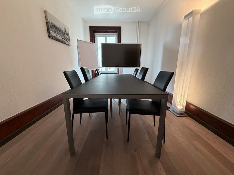 Dining room with a long black dining table and six black chairs, wooden flooring, and a large window with white curtains.