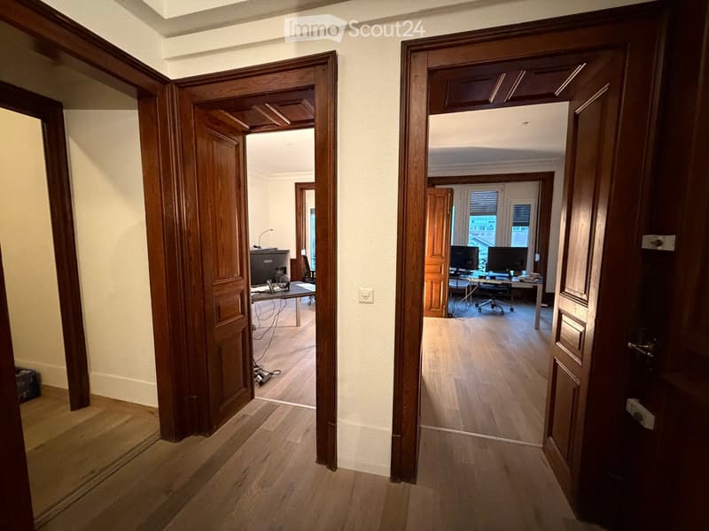 A wide, well-lit hallway with wooden flooring and classic wooden doors leading to furnished rooms, including a workspace with desks and computers.