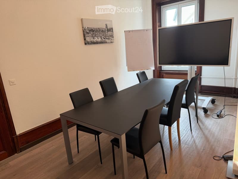 A modern meeting room with a large table, chairs, a screen, and a whiteboard, situated near a window with wooden frames.