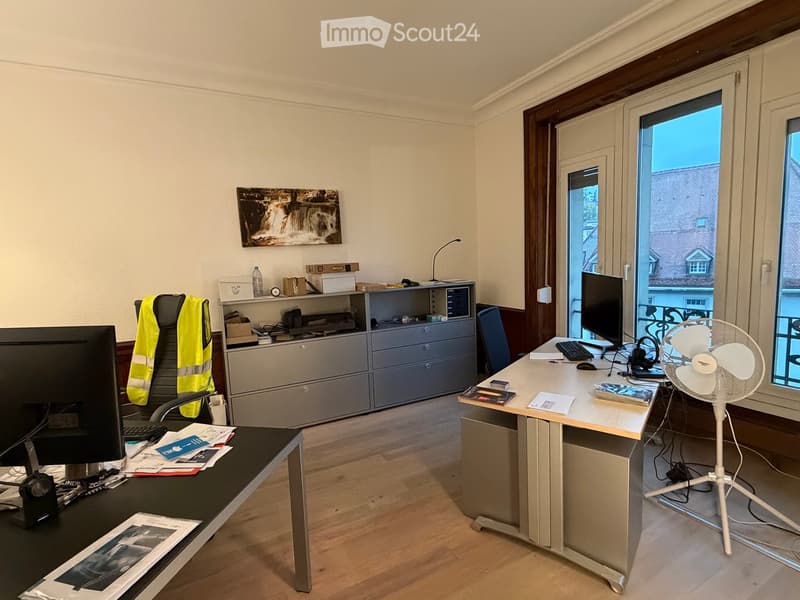 Office space with a wooden floor, a large window, a desk with a computer, a chair, a fan, and a cabinet.