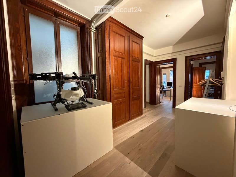 A modern hallway with wooden flooring, large wooden doors, and a small table with a drone on it. There is a visible room with a desk, chair, and screen in the background.