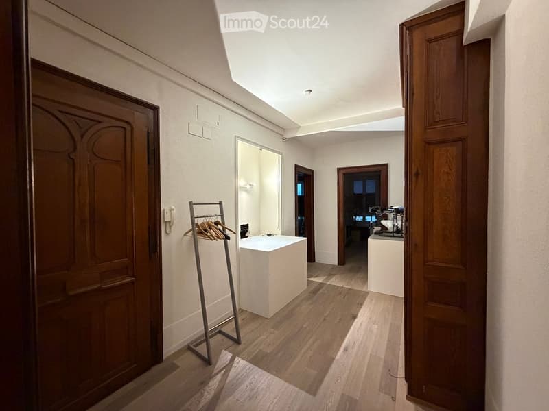 Bright hallway with wooden flooring, white walls, and several doors leading to different rooms. Features include a coat rack with hangers, a small white table, and a mirror on the wall.