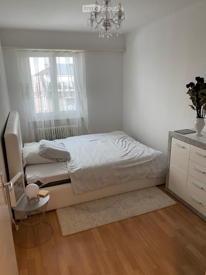 A modern bedroom with a large bed, nightstand, drawer unit, small round table, vase with flowers, chandelier, and picture frame.
