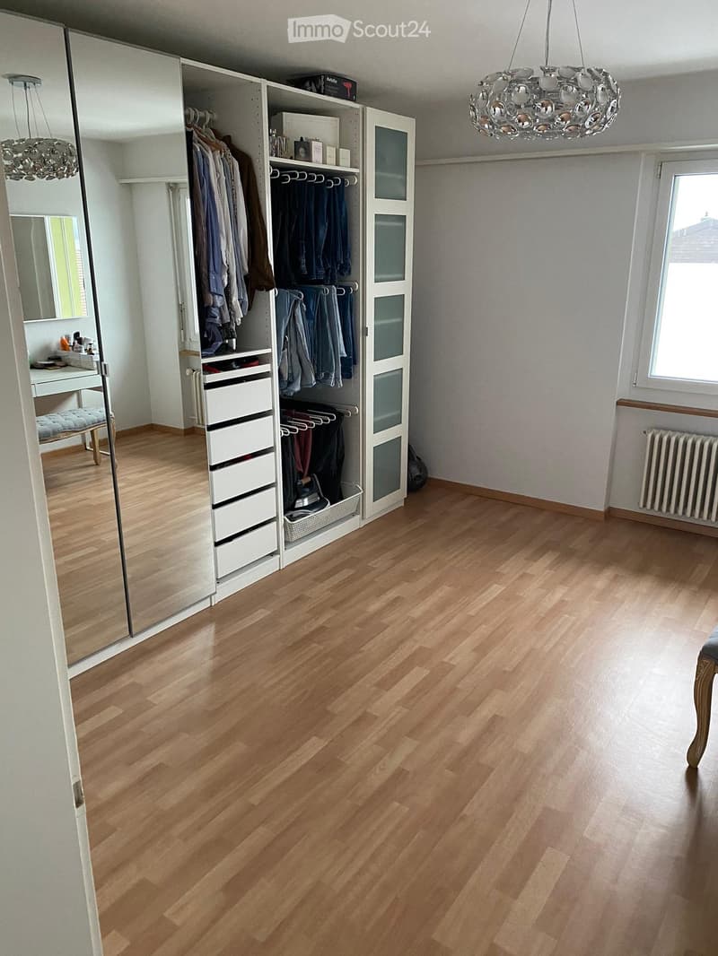 Spacious room with wooden flooring, large built-in wardrobe with hanging space and drawers, a vanity area with a mirror and stool, and a window letting in natural light.