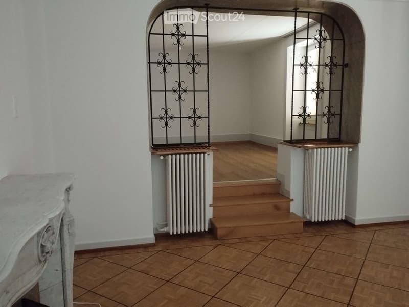 A well-lit hallway with wooden flooring, white walls, and a staircase leading upwards. Features include ornate metal railings, radiators, and a decorative white console table on the left side.