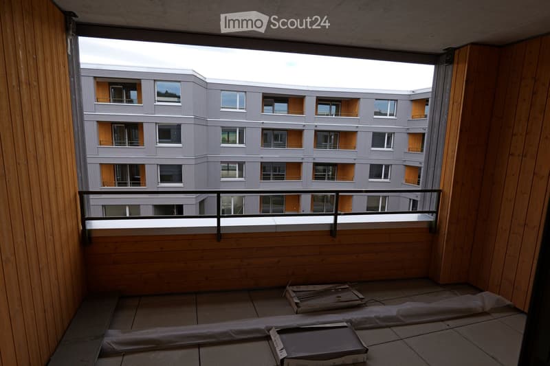 Balcony with wooden flooring and metal railing, overlooking a modern multi-story building with grey and orange accents.