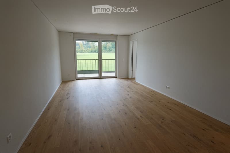 Empty room with light wooden flooring, white walls, and a large sliding glass door leading to a balcony with a view of a green field.