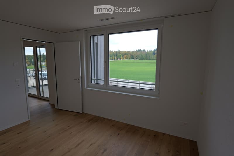 Empty room with wooden flooring, large window overlooking a green field, and a sliding glass door leading to a balcony.
