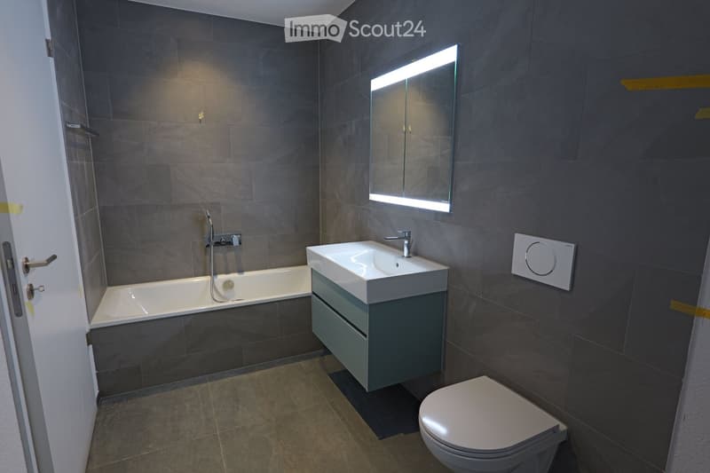 Modern bathroom with dark grey tiles, a bathtub, a sink with a cabinet, a toilet, and a mirror with built-in lighting.