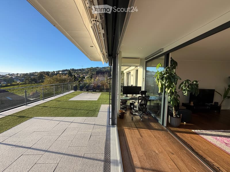 Spacious balcony with wooden flooring, large glass sliding doors, modern furniture including a desk and office chair, potted plants, and a small artificial grass area with lounge chairs.