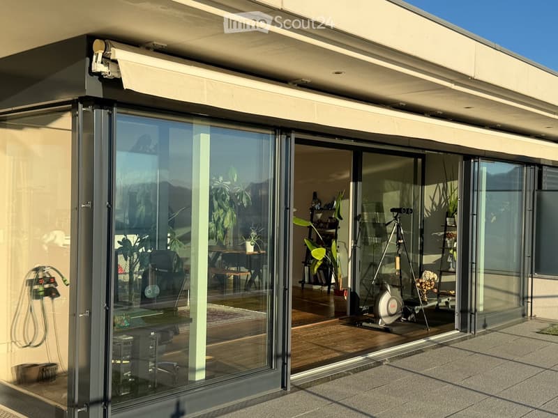 Modern glass-enclosed balcony with retractable awning, large windows, and indoor furniture visible through the glass.